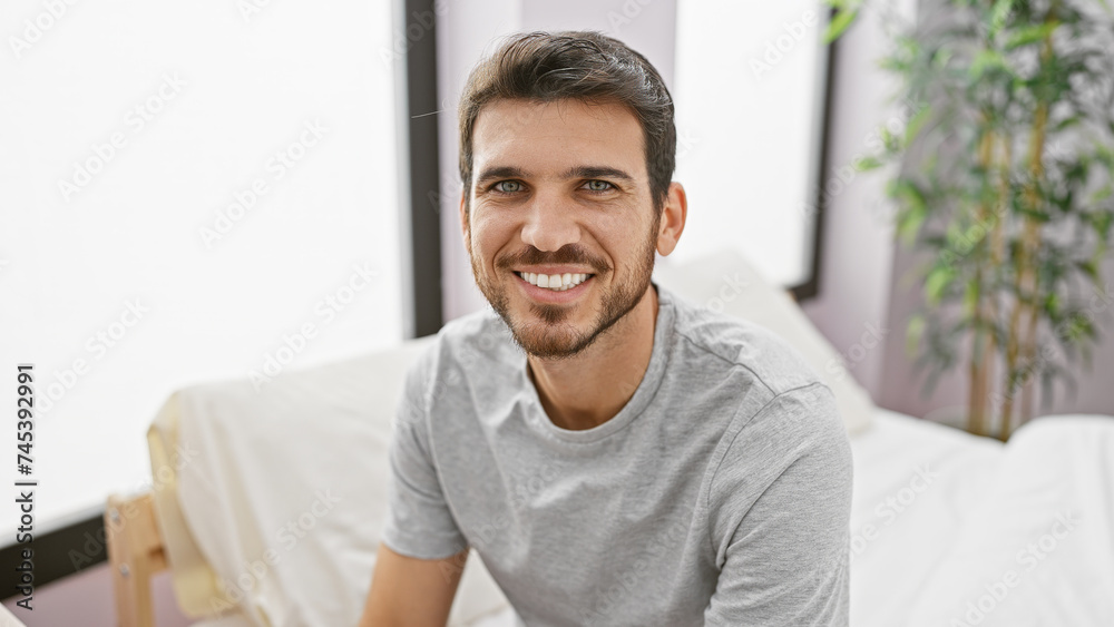 Handsome young hispanic man with a beard smiling in a bright bedroom interior Handsome young hispanic man with a beard smiling in a bright bedroom interior