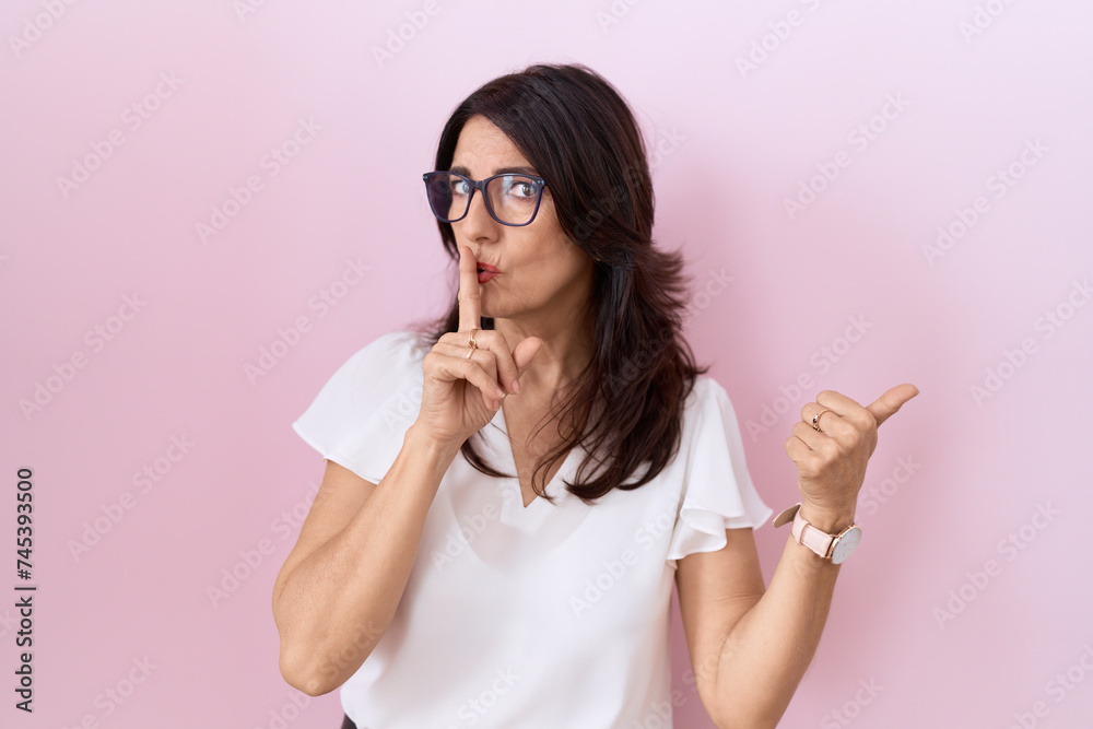 Middle age hispanic woman wearing casual white t shirt and glasses asking to be quiet with finger on lips pointing with hand to the side. silence and secret concept. Middle age hispanic woman wearing casual white t shirt and glasses asking to be quiet with finger on lips pointing with hand to the side. silence and secret concept.