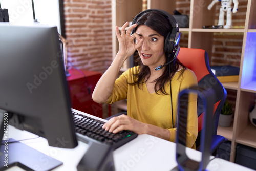 Middle age hispanic woman playing video games using headphones smiling happy doing ok sign with hand on eye looking through fingers