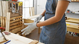 A woman in a denim apron putting on safety gloves in a woodworking workshop
