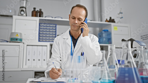 Serious middle-aged man  a scientist absorbed in writing his research report  while talking on a smartphone in the lab.