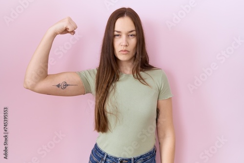 Beautiful brunette woman standing over pink background strong person showing arm muscle, confident and proud of power