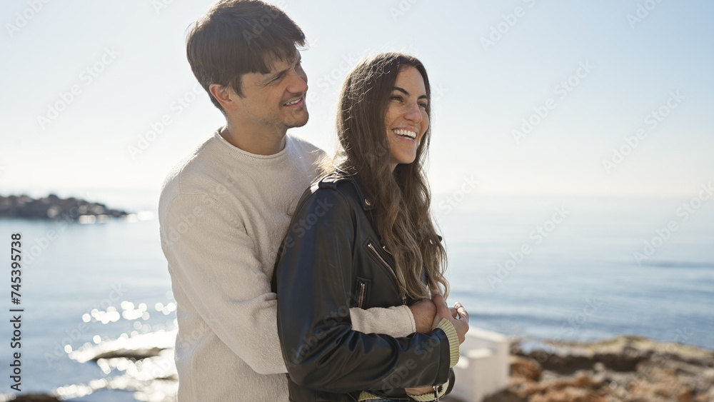 A happy couple embraces beside the serene ocean under clear skies, enjoying a coastal outdoor moment together.