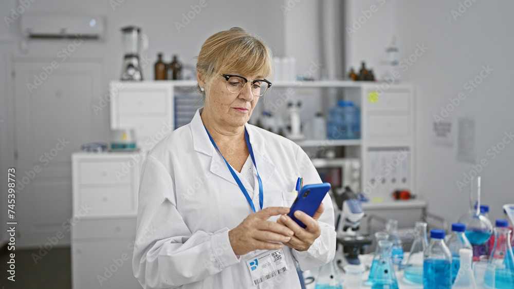 Serious middle age blonde woman scientist engrossed with her smartphone inside a buzzing laboratory, fully immersed in groundbreaking medical research