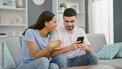 Beautiful couple together in love, confidently smiling, texting on smartphone over coffee on living room sofa at home