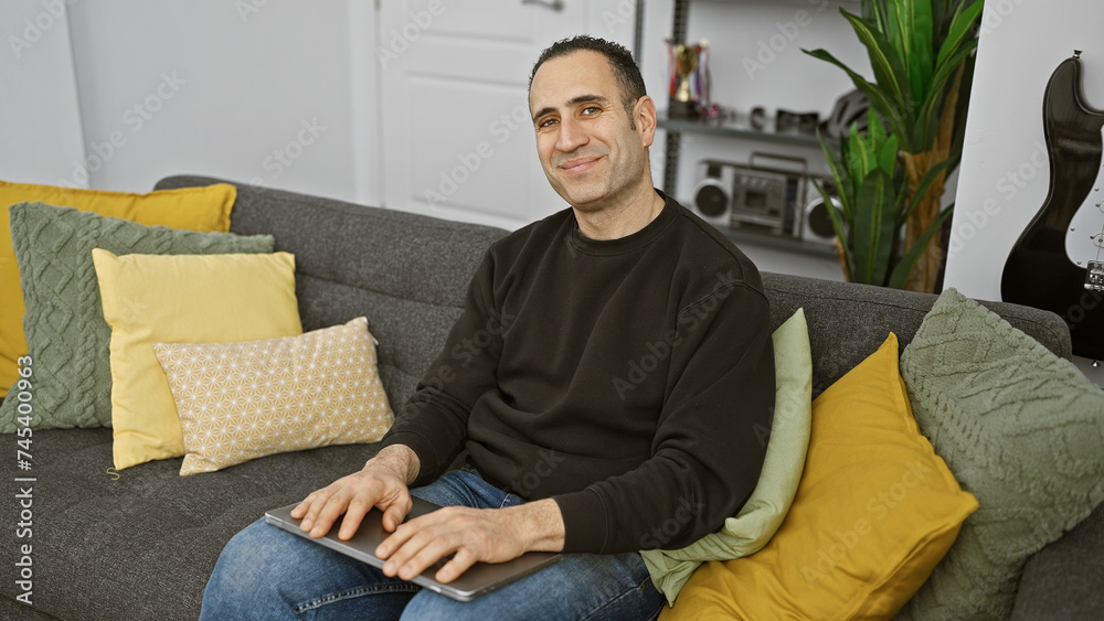 A relaxed hispanic man sits comfortably on a grey sofa in a modern living room, holding a tablet.