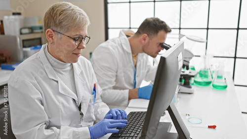 A woman and a man in lab coats work diligently in a bright laboratory, focusing on computer research and scientific analysis.