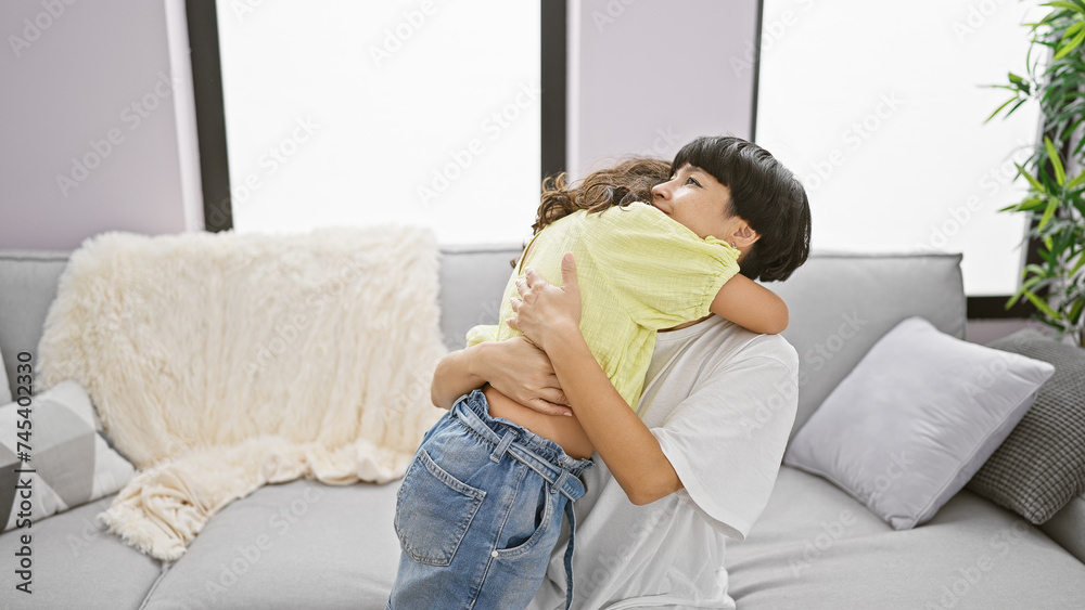 Confident mother and daughter sharing a joyful hug, smiling heartily while sitting on the living room sofa at home – a beautiful expression of family love and happiness Confident mother and daughter sharing a joyful hug, smiling heartily while sitting on the living room sofa at home – a beautiful expression of family love and happiness