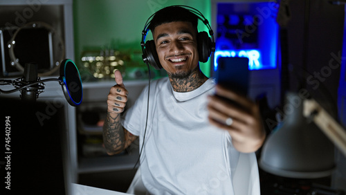 Smiling, tattooed young latin man, a music studio maestro, snapping a vibrant selfie with his smartphone amidst the recording session, donning his headphones. a solo night cap in his artistic haven. photo