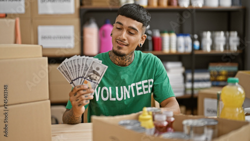 Handsome, tattooed young latin man, smiling confidently, volunteering at the charity center, holding dollars for donations in his hands