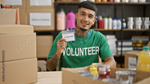 Young, handsome latin man proudly showing off his volunteer id, flashing a confident smile at the heart of a bustling charity center