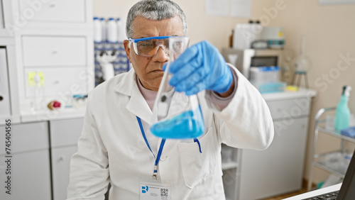 Middle-aged man in lab coat examines contents of a beaker in a laboratory setting, with clinical equipment visible.