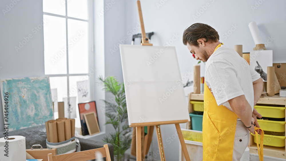 Young hispanic man artist wearing apron standing with serious face at art studio