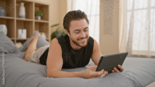 Young hispanic man using touchpad lying on bed at bedroom