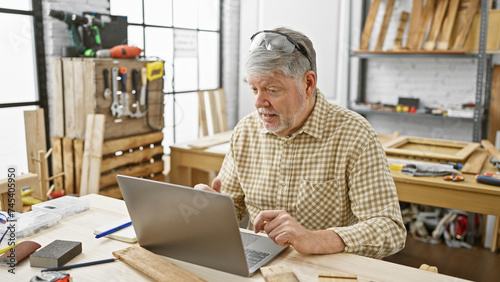 A mature man in a checkered shirt utilizes a laptop in a well-equipped carpentry workshop, surrounded by tools and wood.