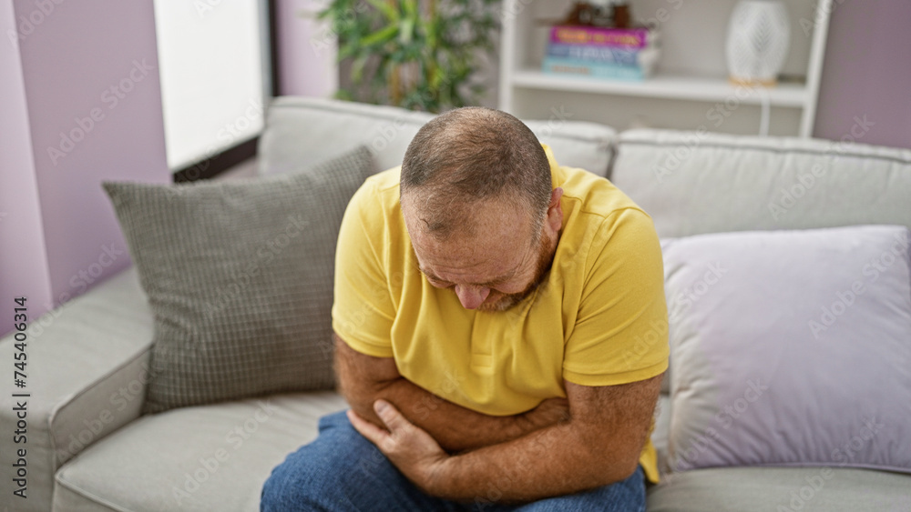 Handsome middle-aged caucasian man sitting on living room sofa at home, touching stomach, seriously worried. adult male suffering from intense stomachache, reflecting illness in his portrait. Handsome middle-aged caucasian man sitting on living room sofa at home, touching stomach, seriously worried. adult male suffering from intense stomachache, reflecting illness in his portrait.