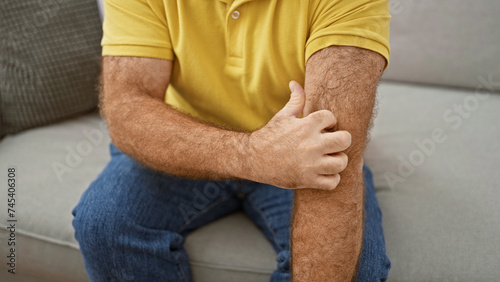 Handsome middle-aged caucasian man sitting on sofa at home, scratching irritated skin on arm. itchy dermatitis rash causing discomfort. health care needed for allergic reaction.