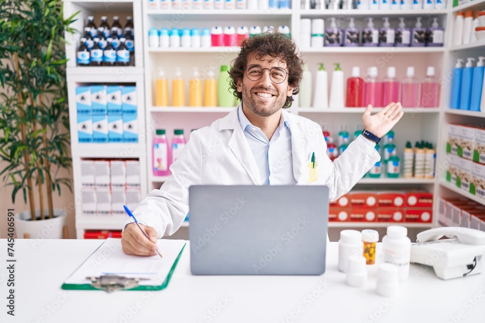 Young hispanic man pharmacist writing on document using laptop at pharmacy