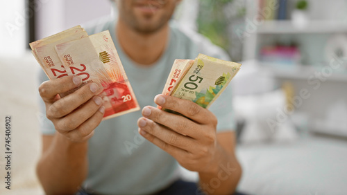 Tableau sur toile Serious young man immersed in counting stack of israeli shekel banknotes on sofa