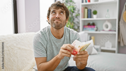 Young man, deep in thought, sits at home on sofa counting israeli shekel banknotes, contemplating his financial wealth and investment strategy