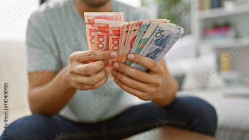 Indoor snapshot, dynamic young man making money count, sitting cozily on sofa at home, banking wealth of yuan banknotes â€“ a peek into chinese economy
