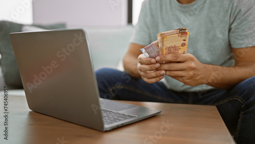 Young man sitting at home, banking online on his laptop, holding wads of rich mexican peso banknotes on his apartment's sofa. photo