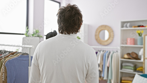 Rear view of a young man in a white shirt standing in a tidy, well-lit dressing room filled with clothes
