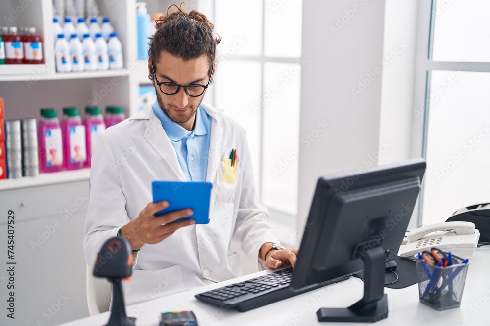 Young hispanic man pharmacist using touchpad and computer, working at pharmacy