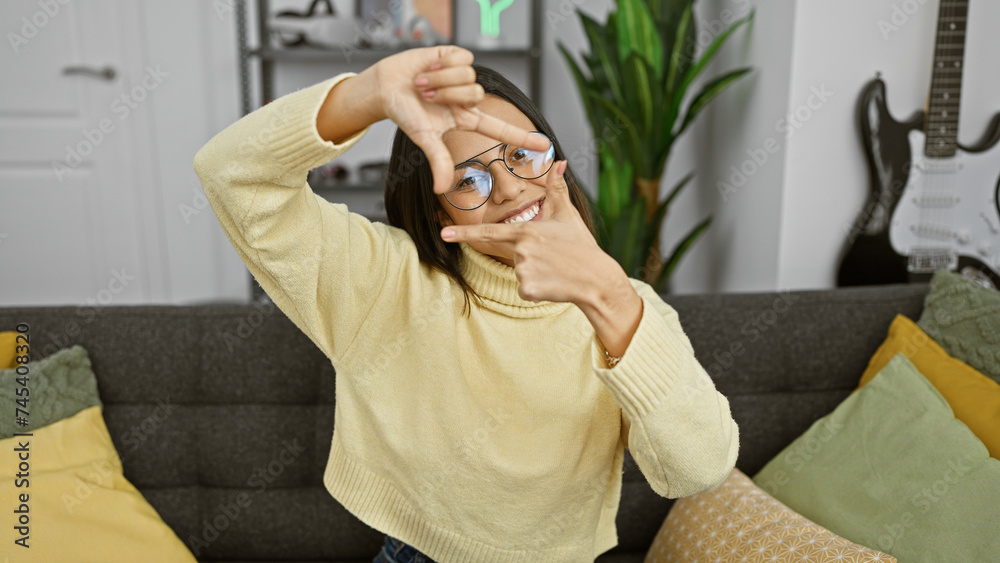 A cheerful young hispanic woman with glasses framing her face with hands, posing indoors in a cozy living room.