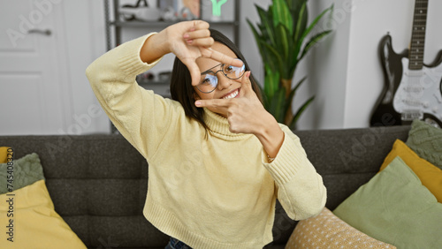 A cheerful young hispanic woman with glasses framing her face with hands, posing indoors in a cozy living room.