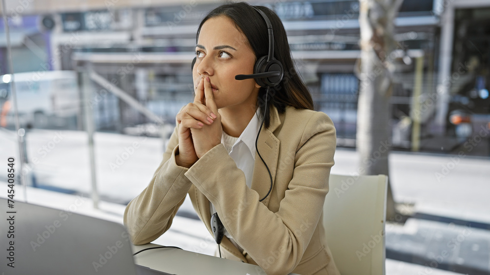 A contemplative hispanic woman in a headset sits at a desk against a blurred office backdrop. A contemplative hispanic woman in a headset sits at a desk against a blurred office backdrop.