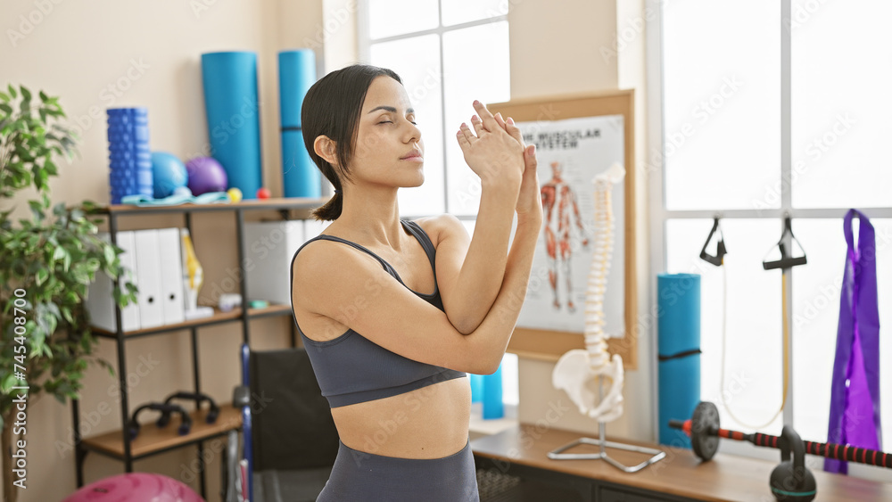 A young hispanic woman in workout attire stretches her arms in a well-equipped physiotherapy clinic room. A young hispanic woman in workout attire stretches her arms in a well-equipped physiotherapy clinic room.