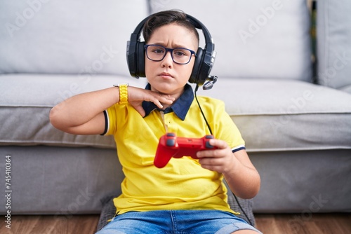 Young hispanic kid playing video game holding controller wearing headphones cutting throat with hand as knife, threaten aggression with furious violence