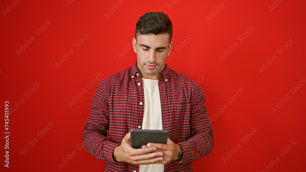 Cool young hispanic man absorbed in his touchpad, standing casual over red isolated background. portrait of attractive male with serious expression, immersed in online world on his device. Cool young hispanic man absorbed in his touchpad, standing casual over red isolated background. portrait of attractive male with serious expression, immersed in online world on his device.
