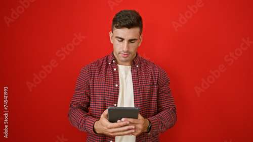 Cool young hispanic man absorbed in his touchpad, standing casual over red isolated background. portrait of attractive male with serious expression, immersed in online world on his device. © Krakenimages.com