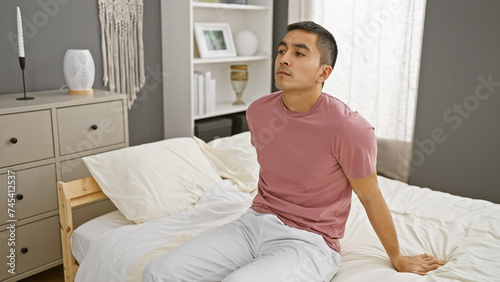 Handsome young hispanic man sitting thoughtfully on a bed in a modern bedroom interior.