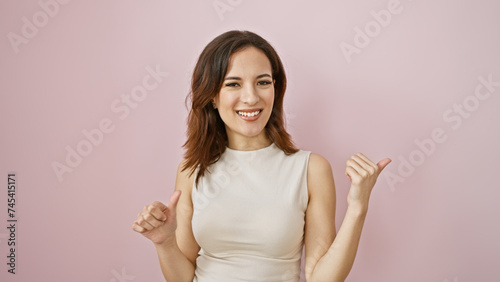 A young hispanic woman smiles charmingly while pointing sideways against a soft pink background, evoking positivity and beauty.