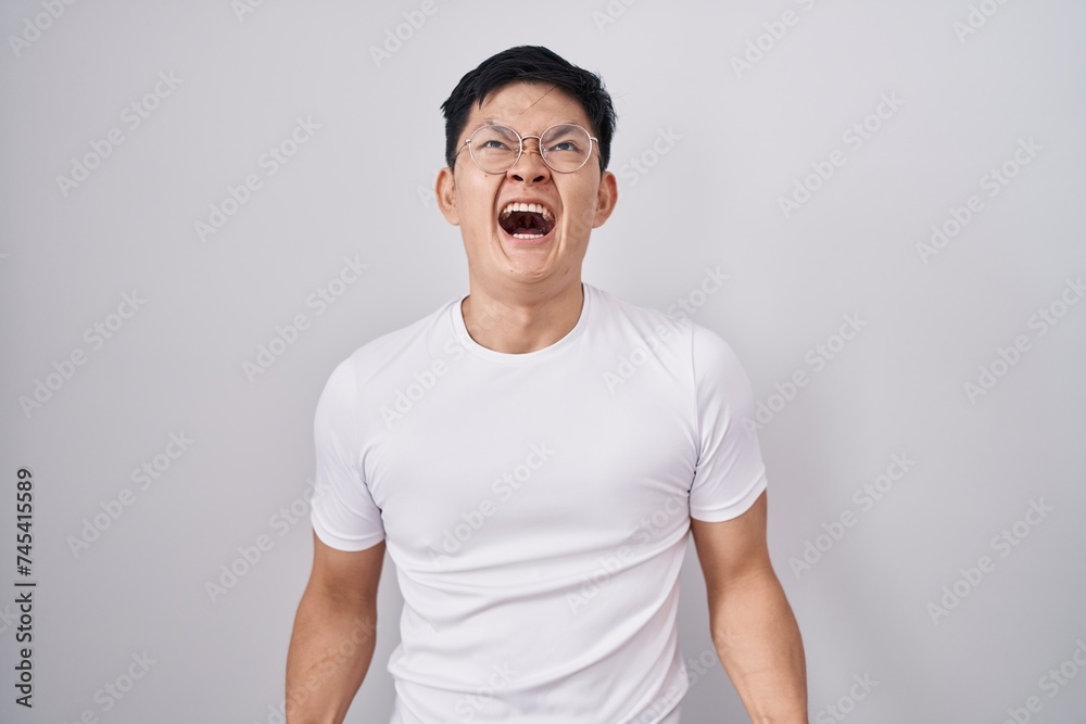 Young asian man standing over white background angry and mad screaming frustrated and furious, shouting with anger. rage and aggressive concept. Young asian man standing over white background angry and mad screaming frustrated and furious, shouting with anger. rage and aggressive concept.