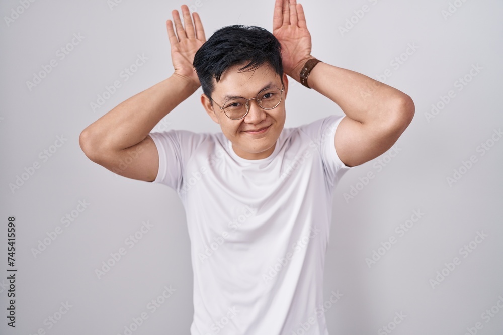 Young asian man standing over white background doing bunny ears gesture with hands palms looking cynical and skeptical. easter rabbit concept.