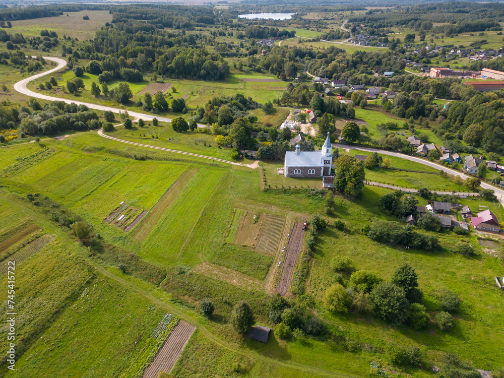 Fototapeta premium Top view of a small village in the middle of a green field and forest. An old church on a hill in the center of the city. Aerial view of the farm field and the natural landscape.