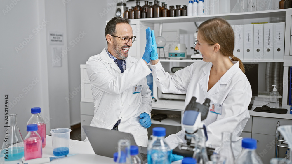 Two thrilled scientists high five at lab, celebrating discovery on laptop. man and woman, partners in science, beam amid a backdrop of test tubes and microscopes.