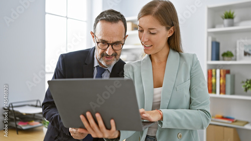 Two confident business workers smiling as they work together on a laptop, standing in the heart of a bustling office environment