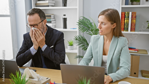 Фотография Two business workers sneezing together at office while hard at work on a laptop, a clear expression of office flu affliction indoors