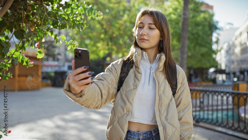 A young caucasian woman takes a selfie outdoors on a sunny city street, surrounded by greenery.