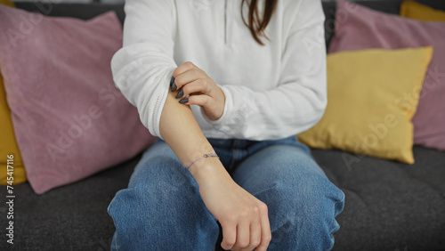 A young woman scratches her arm while sitting comfortably in a modern living room, adding a personal touch to the cozy home setting.