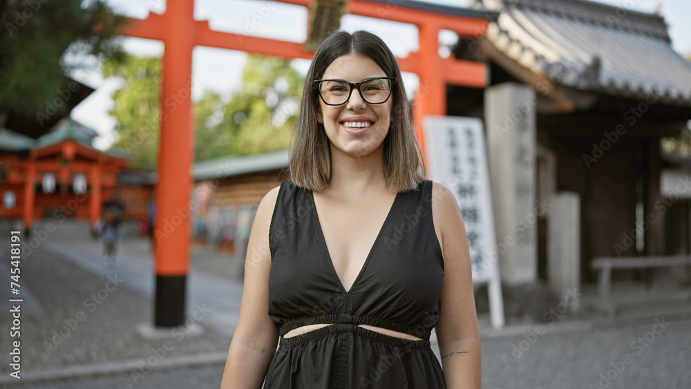 Beautiful hispanic woman with glasses poses at fushimi inari-taisha, joy radiating from her successful smile, confidence stands strikingly in kyoto's grand shrine