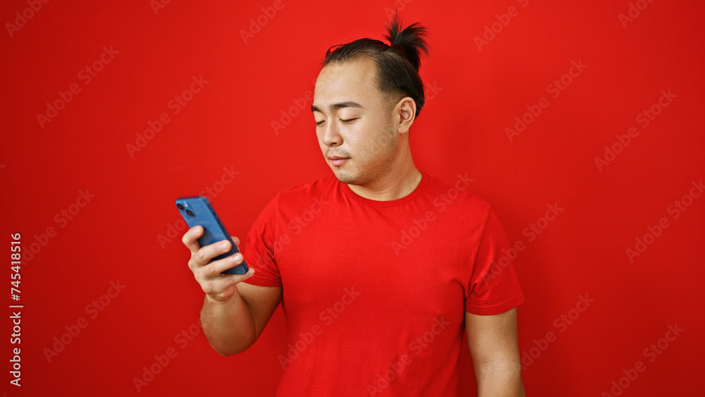 Handsome young chinese man, in relaxed concentration, uses his phone, tapping into the digital world over an isolated red background Handsome young chinese man, in relaxed concentration, uses his phone, tapping into the digital world over an isolated red background