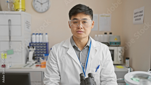 A young asian man wearing safety goggles and a lab coat stands confidently in a clinical laboratory setting.