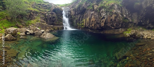 A small waterfall cascading into the center of a lake, creating ripples on the waters surface. The waterfall is surrounded by lush greenery and rocks.