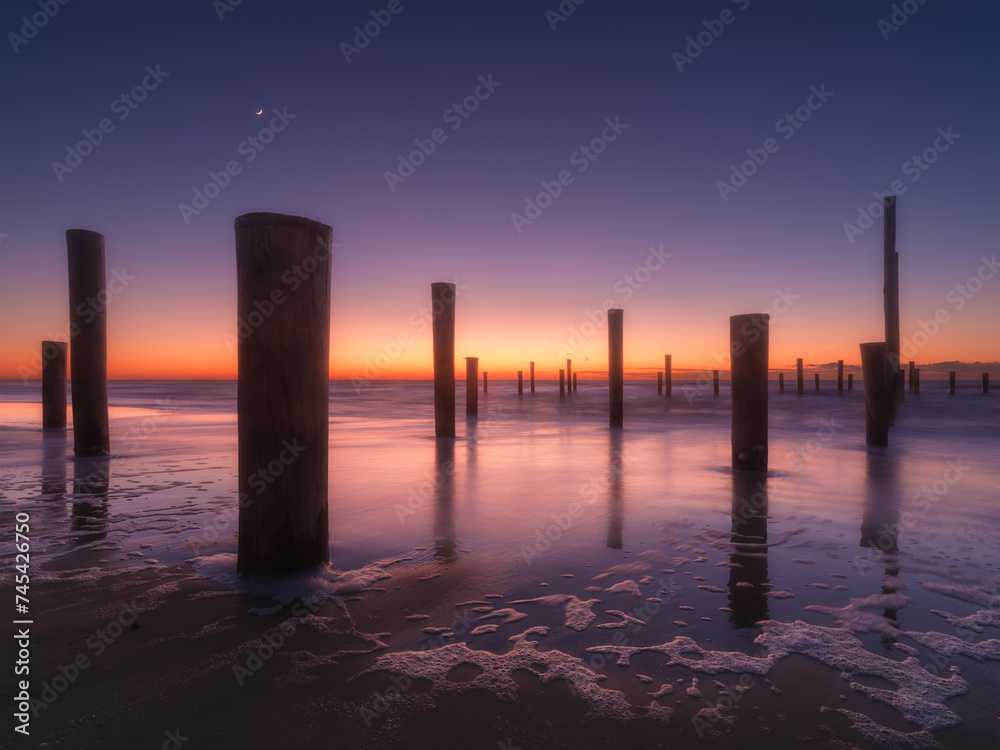 A seascape during sunset. Pillars on the seashore. Bright sky during sunset. Reflections on the seashore. A sandy beach at low tide. Travel image.